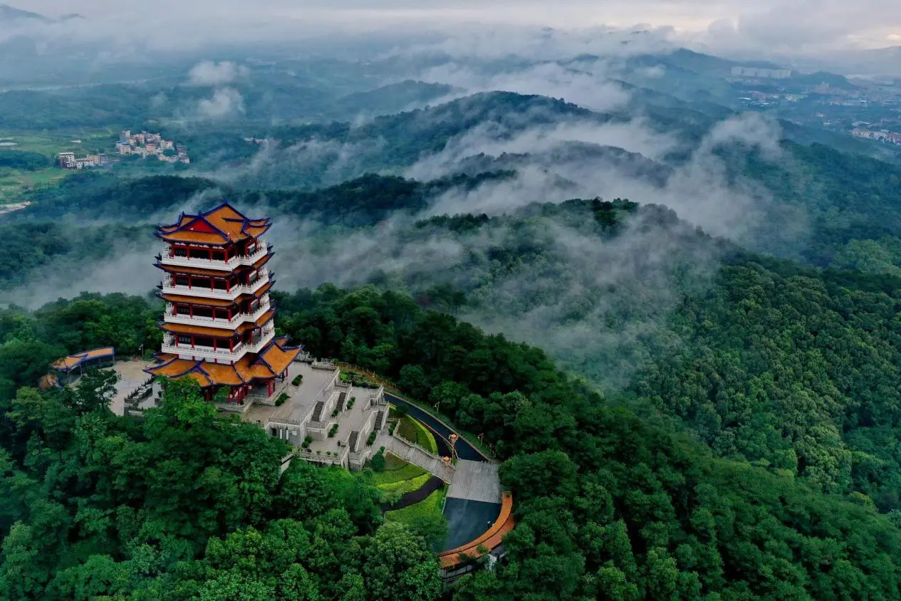 Drone shot of Shaoyang Tower perched on a forested mountain peak surrounded by clouds and mist, with a distant village visible in the valley — a cultural landmark of Shaoguan, Guangdong